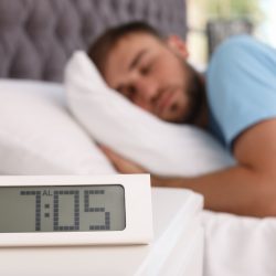 Alarm clock on table and young man sleeping in bed at home