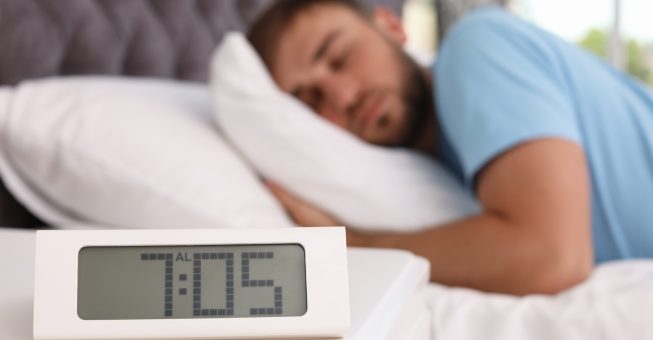 Alarm clock on table and young man sleeping in bed at home