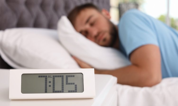 Alarm clock on table and young man sleeping in bed at home