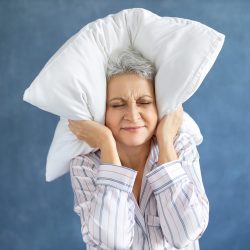 Annoyed dissatisfied mature woman with gray hair grimacing having sleepless night because of loud music, snoring husband, covering ears with pillow, having angry irritated facial expression