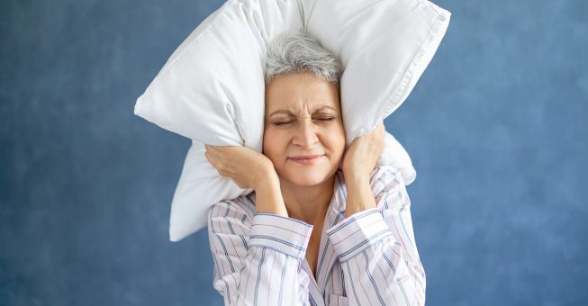 Annoyed dissatisfied mature woman with gray hair grimacing having sleepless night because of loud music, snoring husband, covering ears with pillow, having angry irritated facial expression