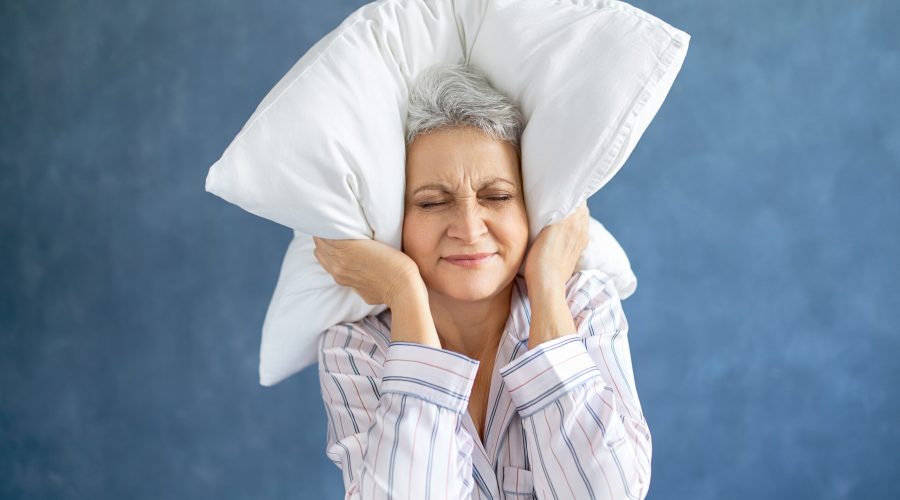 Annoyed dissatisfied mature woman with gray hair grimacing having sleepless night because of loud music, snoring husband, covering ears with pillow, having angry irritated facial expression