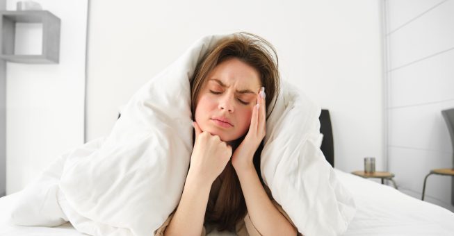 Portrait of woman rolled in blankets in bed, frowning and feeling unwell, has hangover, headache in morning, resting at home.