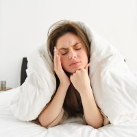 Portrait of woman rolled in blankets in bed, frowning and feeling unwell, has hangover, headache in morning, resting at home.