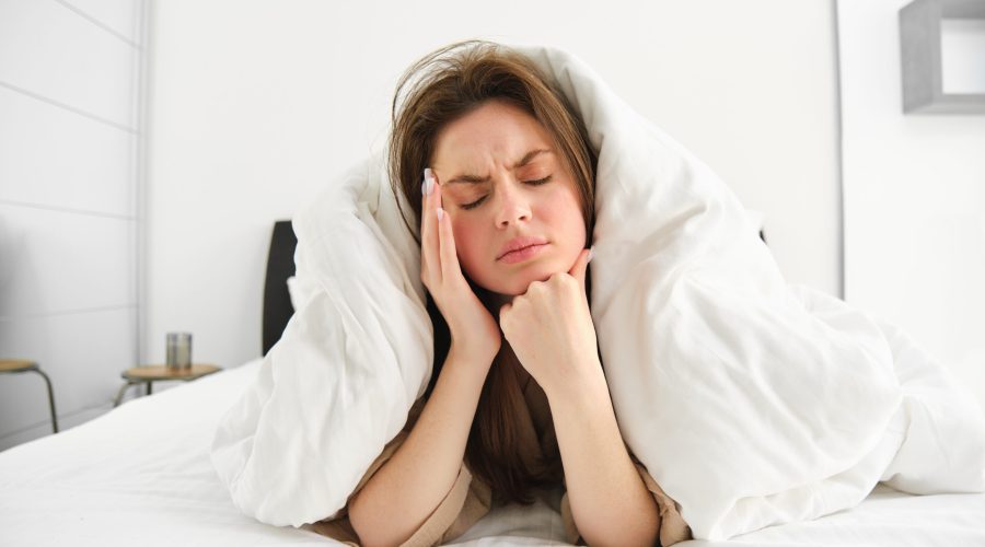 Portrait of woman rolled in blankets in bed, frowning and feeling unwell, has hangover, headache in morning, resting at home.