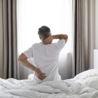 Unrecognizable mature grey-haired man wearing white pajamas sittting on bed at home in the morning, touching lower back and neck, suffering from muscle strain after waking up, copy space