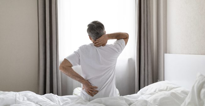 Unrecognizable mature grey-haired man wearing white pajamas sittting on bed at home in the morning, touching lower back and neck, suffering from muscle strain after waking up, copy space