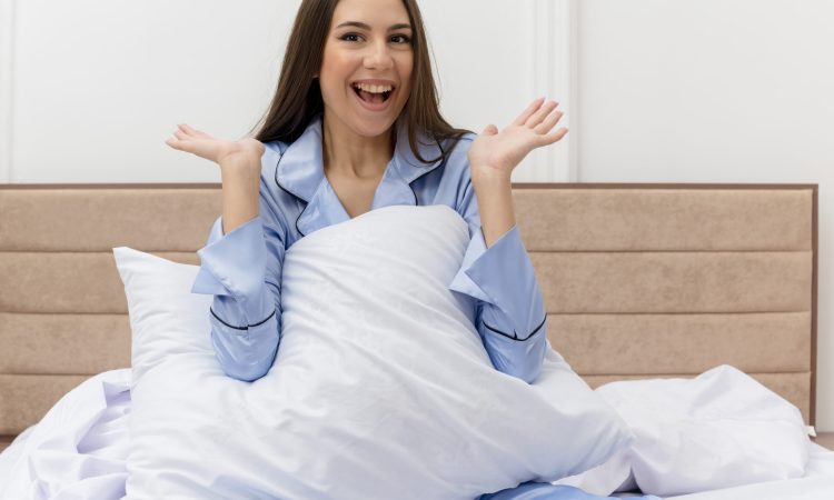 young beautiful woman in blue pajamas sitting on bed with pillows resting looking at camera happy and positive smiling enjoying weekend in bedroom interior on light background