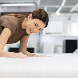 Young beautiful woman choosing mattress at retail furniture store. Housewife touching softness test buying bedding for family. Female checking quality shopping at mattress store hall.