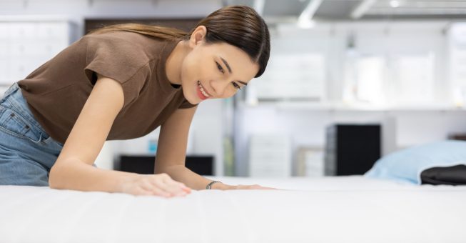 Young beautiful woman choosing mattress at retail furniture store. Housewife touching softness test buying bedding for family. Female checking quality shopping at mattress store hall.