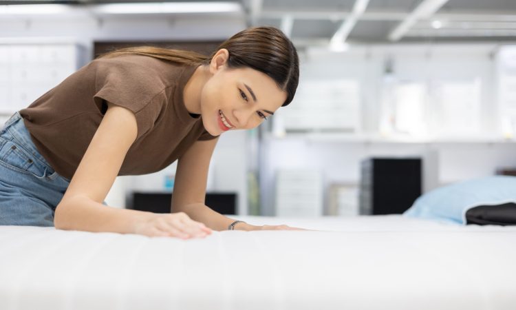 Young beautiful woman choosing mattress at retail furniture store. Housewife touching softness test buying bedding for family. Female checking quality shopping at mattress store hall.