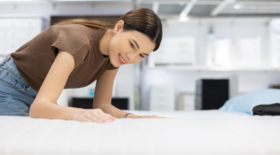 Young beautiful woman choosing mattress at retail furniture store. Housewife touching softness test buying bedding for family. Female checking quality shopping at mattress store hall.