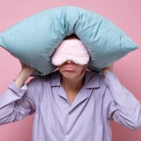 Young caucasian woman wearing sleep mask during sleeping holding a pillow. Studio shot on pink wall.