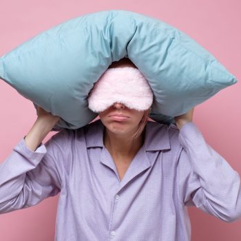 Young caucasian woman wearing sleep mask during sleeping holding a pillow. Studio shot on pink wall.