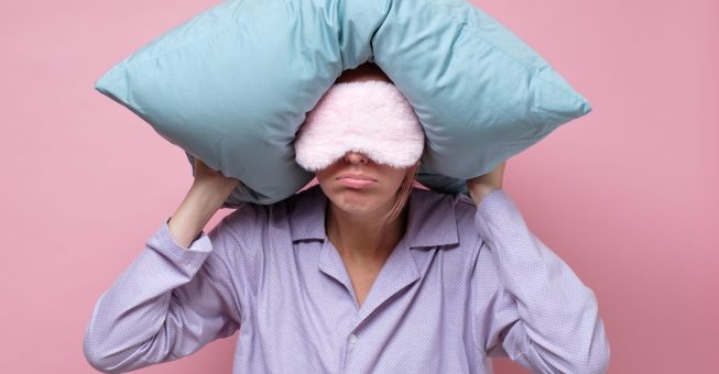 Young caucasian woman wearing sleep mask during sleeping holding a pillow. Studio shot on pink wall.