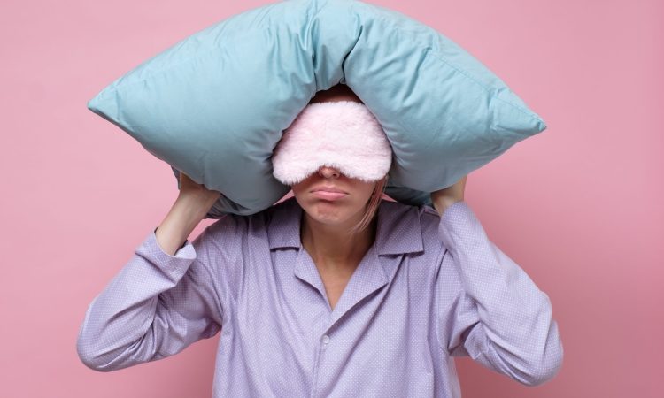 Young caucasian woman wearing sleep mask during sleeping holding a pillow. Studio shot on pink wall.